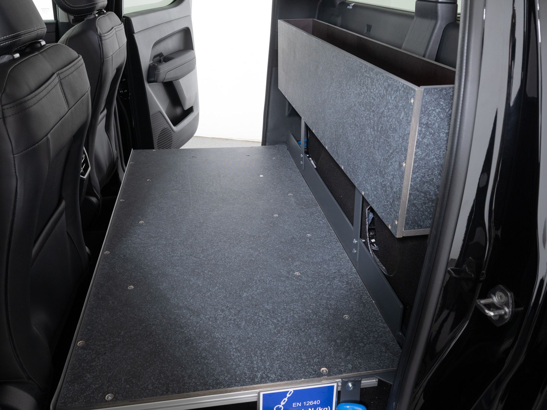 Der Innenraum eines Fahrzeugs, das eine umgeklappte Liegefläche zeigt. Die Liegefläche ist schwarz und aus einem festen Material. Am unteren Ende der Liegefläche ist ein blauer Aufkleber angebracht.