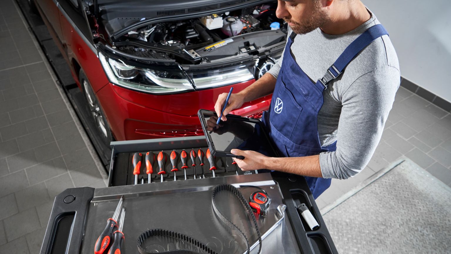 Ein Volkswagen Mechaniker in blauer Arbeitskleidung steht vor einem roten Volkswagen mit geöffneter Motorhaube und benutzt ein Tablet. Auf einem Rollwagen vor ihm liegen verschiedene Werkzeuge, darunter Schraubendreher, Zangen und ein Zahnriemen.