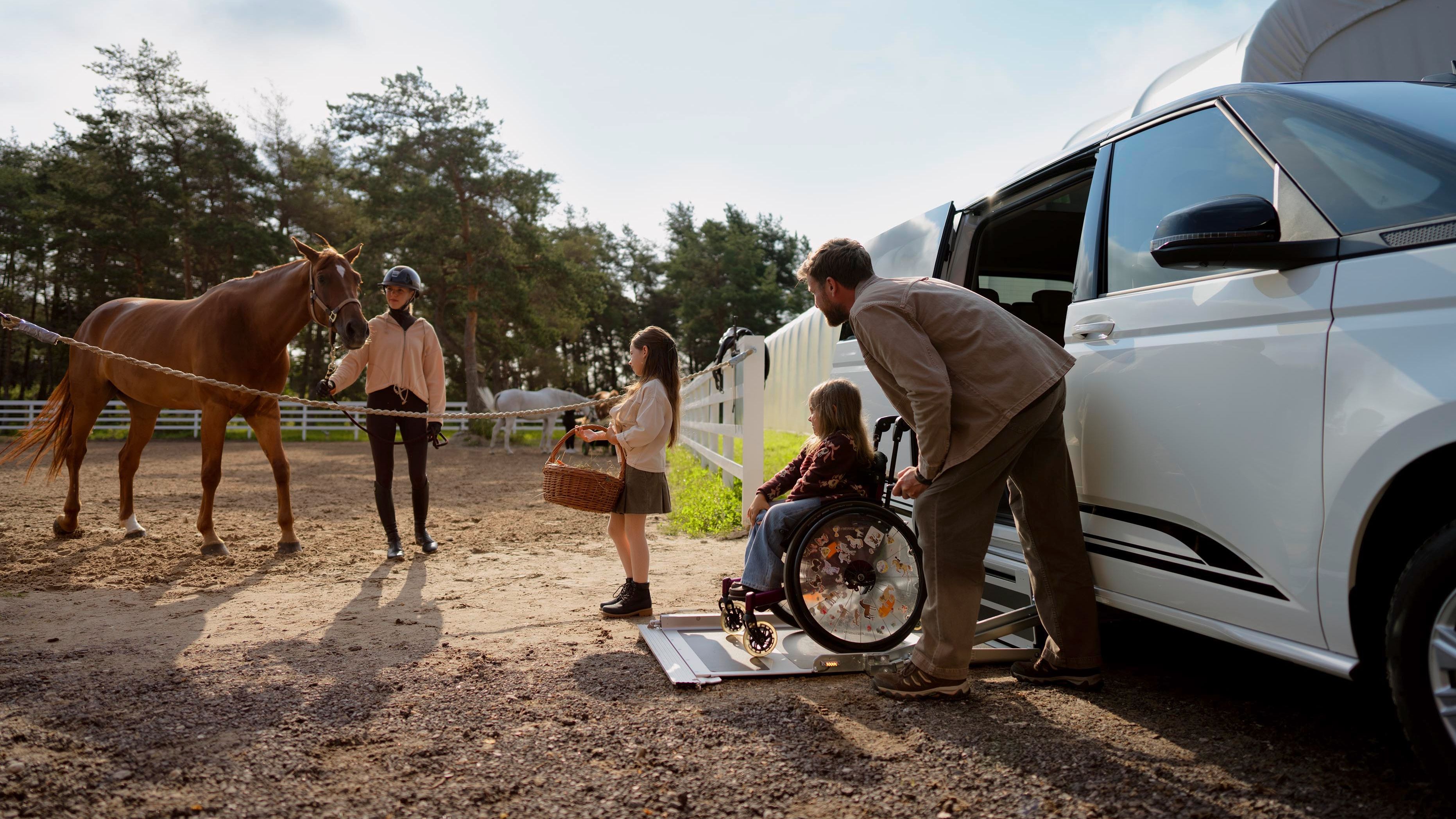 Ein Mann hilft einer Person im Rollstuhl, über eine Rampe aus einem weißen Fahrzeug zu steigen. Eine Frau und ein Kind stehen neben dem Fahrzeug und halten einen Picknickkorb. Im Hintergrund ist ein braunes Pferd zu sehen.