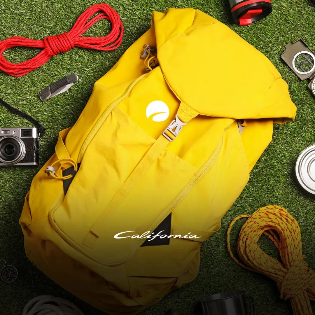 Ein gelber Wanderrucksack liegt auf Gras, umgeben von verschiedenen Camping- und Outdoor-Ausrüstungsgegenständen wie einem Seil, einer Kamera, einer Taschenlampe und Werkzeugen.