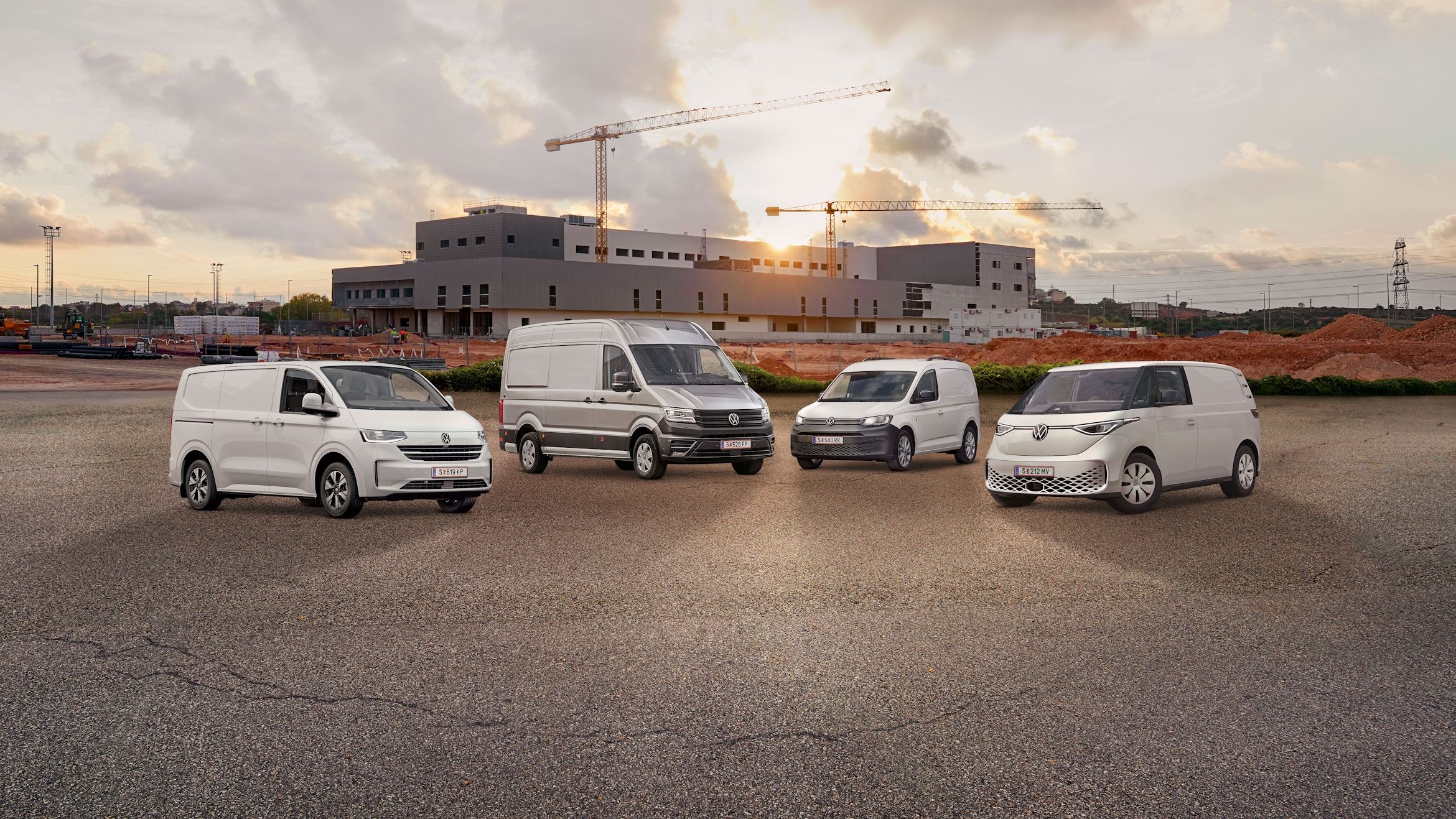 Vier weiße Transporter von Volkswagen Nutzfahrzeuge stehen nebeneinander auf einem leeren Parkplatz vor einem unfertigen Gebäude. Der Himmel ist bewölkt mit einigen Sonnenstrahlen. Ein Baukran ist im Hintergrund sichtbar.