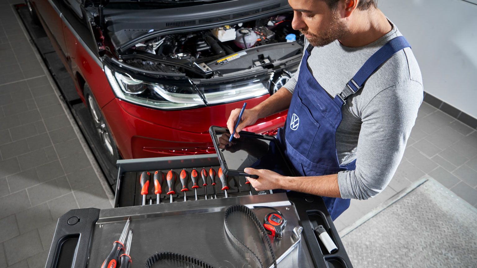 Ein Volkswagen Mechaniker in blauer Arbeitskleidung steht vor einem roten Volkswagen mit geöffneter Motorhaube und benutzt ein Tablet. Auf einem Rollwagen vor ihm liegen verschiedene Werkzeuge, darunter Schraubendreher, Zangen und ein Zahnriemen.