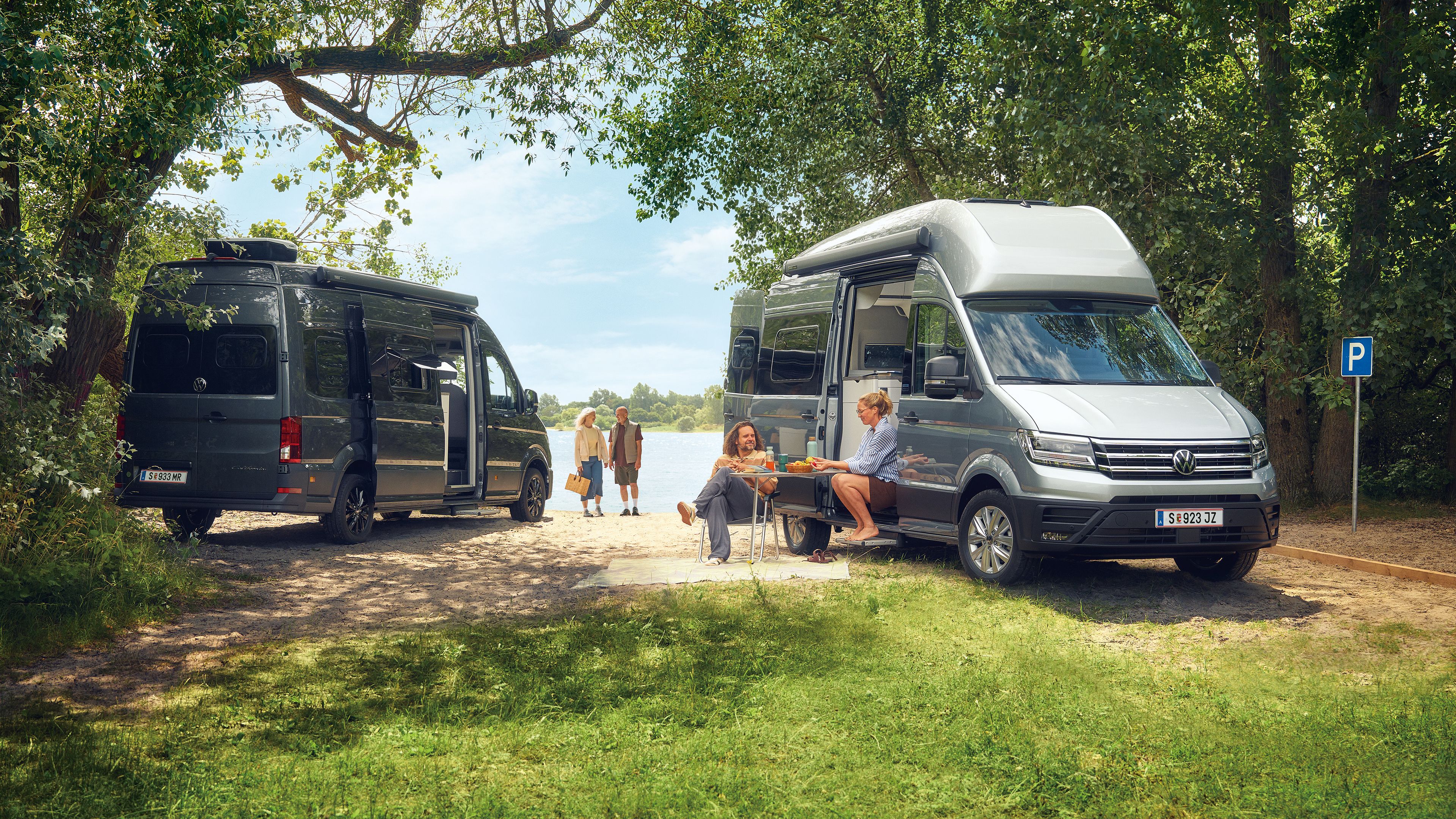Zwei VW Grand California stehen auf einer grasbewachsenen Fläche in der Nähe eines Sees. Es gibt mehrere Personen; eine sitzt an einem Picknicktisch und zwei weitere gehen auf das Wasser zu. Die Szene ist von Bäumen und Grün umgeben.