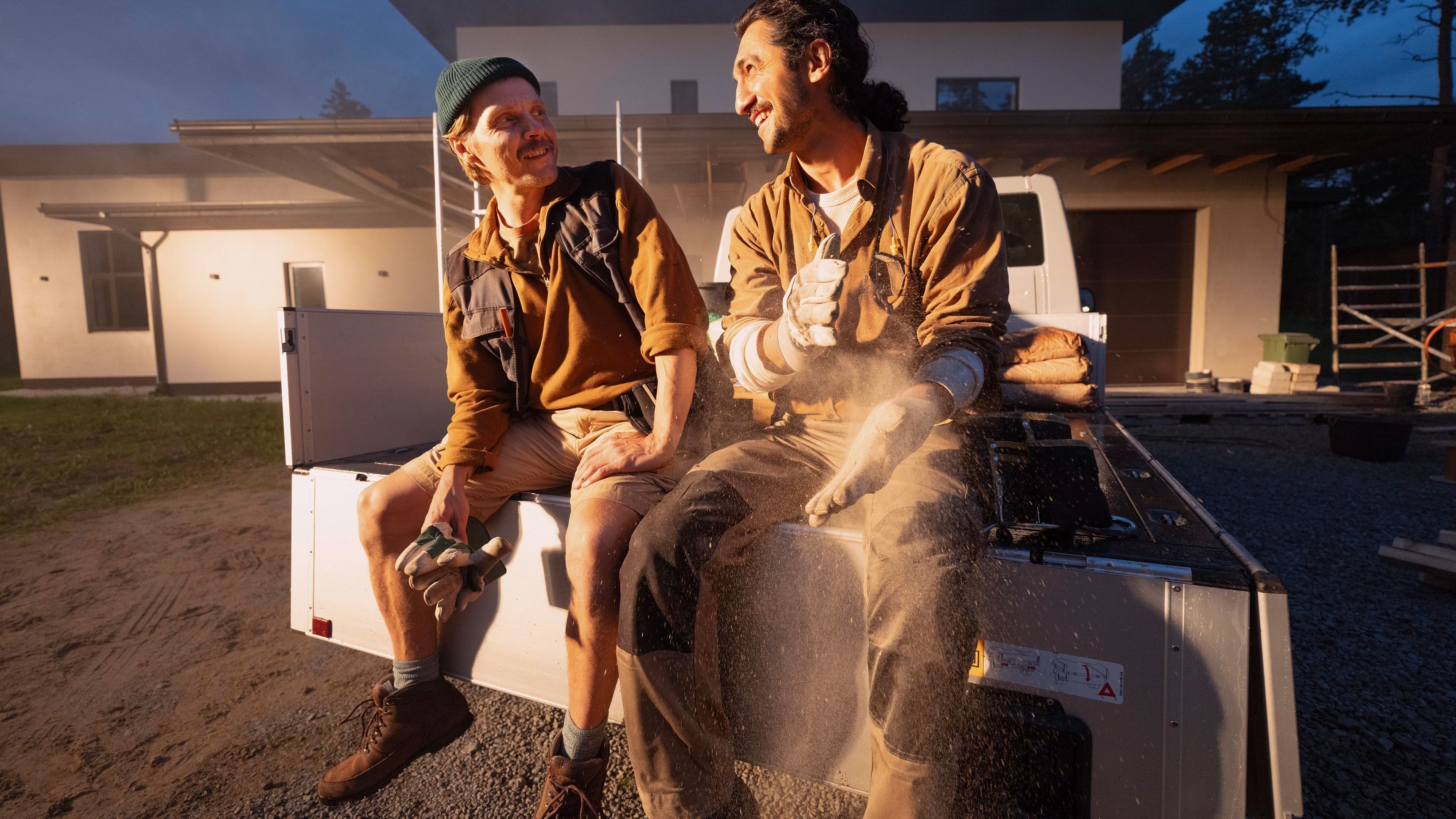Zwei Personen sitzen auf der Ladefläche eines Pick-up Trucks vor einem modernen Gebäude. Sie tragen Arbeitskleidung und schützen sich vor Staub. Die Umgebung scheint eine Baustelle zu sein.