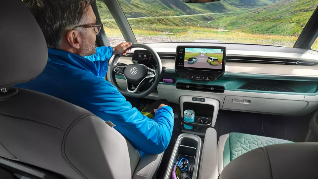 Ein Mann sitzt in einem VW ID. Buzz und fährt durch eine bergige Landschaft. Das Auto hat einen großen Bildschirm, der Navigationsdaten anzeigt. Die Inneneinrichtung ist elegant und technologisch fortgeschritten.