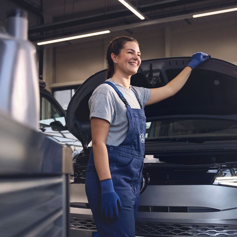 Eine Volkswagen Mechanikerin in blauer Arbeitskleidung steht in einer Volkswagen Werkstatt und inspiziert den Motorraum eines offenen Volkswagen. Sie trägt Handschuhe und stützt sich mit einer Hand auf die Motorhaube.