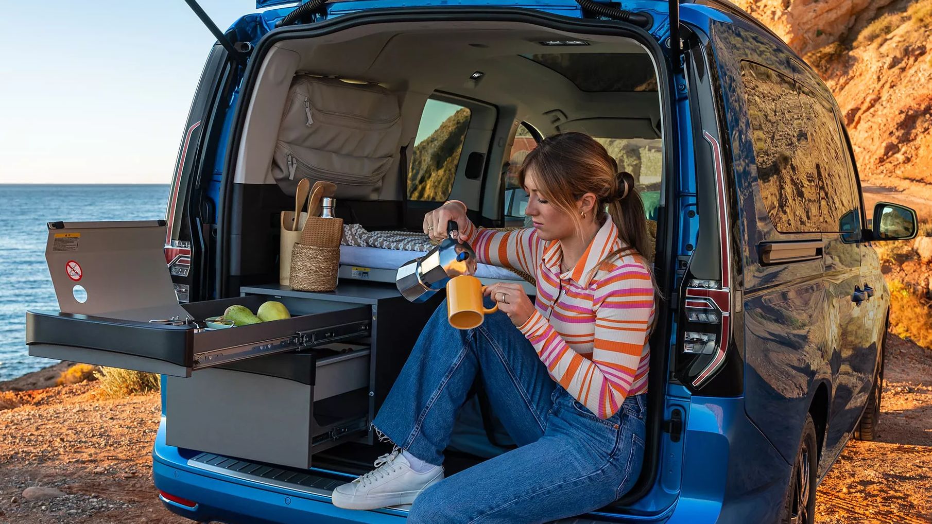 Eine Frau sitzt bei geöffneter Heckklappe in ihrem VW Caddy California, der am Meer parkt